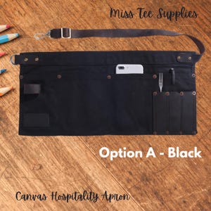 Puede incluir: Delantal de hospitalidad de lona negra con una correa de cuero, un bolsillo para teléfono y un bolsillo para herramientas. El delantal tiene remaches metálicos y una solapa de cuero. "Miss Tee Supplies" y "Opción A - Negro" están impresos en el delantal.