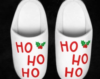 Può includere: Coppia di pantofole bianche con il testo "HO HO HO" in rosso e un ramo di agrifoglio verde con bacche rosse.