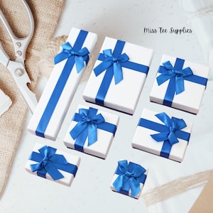 Pode incluir: Conjunto de caixas de presente brancas com laços de fita azul. As caixas são de vários tamanhos e estão dispostas sobre uma superfície branca. Miss Tee Supplies está escrito no canto superior direito da imagem.