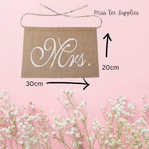Op de afbeelding: Een jute banner met het woord "Mrs." geschreven in witte, cursieve letters. De banner is 20 cm hoog en 30 cm breed.