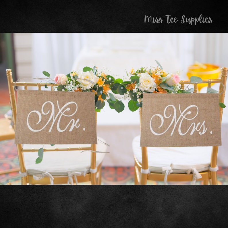 Op de afbeelding: Twee gouden stoelen met witte kussens en juteborden met de tekst "Mr." en "Mrs.". De stoelen zijn versierd met een krans van witte en oranje bloemen en groen.