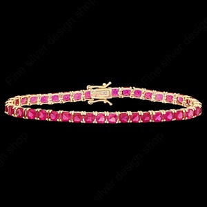 May include: A gold-toned bracelet featuring a row of square-cut, vibrant red gemstones. The bracelet has a secure clasp and is set against a black background, highlighting the jewelry's sparkle and design.