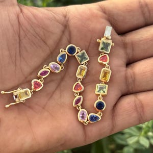 May include: A gold bracelet with a variety of colorful gemstones, including blue, green, red, pink, and yellow. The bracelet is made up of different shapes, including squares, rectangles, and hearts.