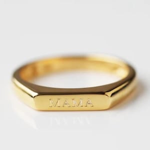 Op de afbeelding: Een goudkleurige ring met het woord "MAMA" gegraveerd op de voorkant. De ring heeft een rechthoekige vorm met licht afgeronde randen. De ring staat tegen een witte achtergrond.