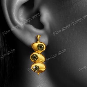 May include: Gold-colored earrings with three olive-green gemstones. The earrings have a unique, elongated design, with each gemstone set within a rounded, gold-toned shape. The background is a black and white image of a person's ear.