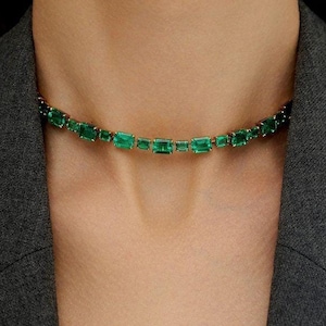 Puede incluir: Un collar de cadena de oro con piedras rectangulares de color verde esmeralda. El collar está en un modelo que lleva una chaqueta negra.