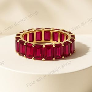 Puede incluir: Un anillo dorado con una banda de piedras preciosas rectangulares de color rojo intenso. El anillo se exhibe sobre un pedestal blanco y circular, sobre un fondo neutro. Las piedras preciosas están muy juntas, creando una banda continua de color.