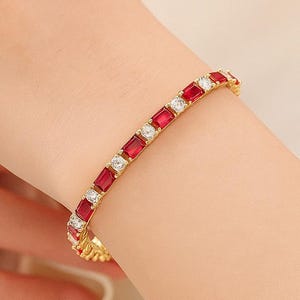 Könnte beinhalten: Ein goldenes Armband mit abwechselnd rechteckigen roten und klaren Edelsteinen. Das Armband wird am Handgelenk getragen, wodurch das Design und der Farbkontrast hervorgehoben werden. Die Edelsteine sind in einem goldenen Band gefasst.