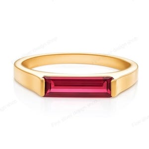 May include: A gold ring featuring a rectangular, ruby-coloured gemstone. The ring has a simple, modern design with a polished finish. The gemstone is set horizontally in the centre of the band, creating a sleek and elegant look. The ring is likely made of gold.