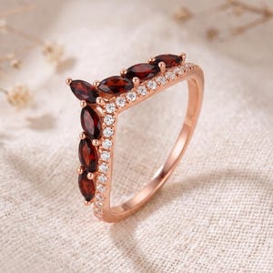 May include: A rose gold V-shaped ring featuring a row of marquise-cut deep red garnet gemstones and a parallel row of small round white accent stones. The band is crafted from a polished rose-colored metal, creating a chevron silhouette.