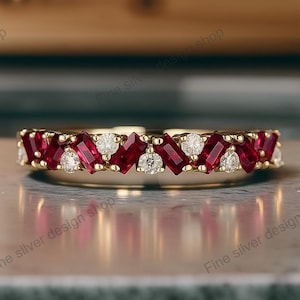 Puede incluir: Un anillo de banda de oro con piedras preciosas rojas y blancas alternas. Las piedras rojas son rectangulares, mientras que las blancas son redondas y triangulares. El anillo está sobre una superficie reflectante.