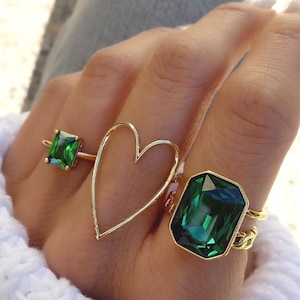 May include: Three gold rings with green gemstones. The rings feature a heart shape, a square emerald cut stone, and a larger emerald cut stone.