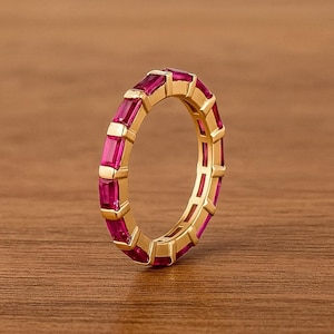 May include: A gold-colored ring with a continuous band of rectangular, magenta-colored gemstones. The ring is set on a wooden surface, showcasing the contrast between the warm gold and vibrant gemstones. The gemstones are evenly spaced around the ring.