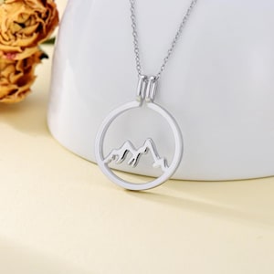 Puede incluir: Un collar de plata con un colgante circular que presenta un diseño recortado de montañas.