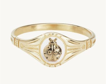 May include: Gold-tone ring featuring a detailed ladybug design set within a white oval. The ring has a smooth band with decorative accents on the shoulders. A charming piece of jewelry.