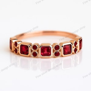 Puede incluir: Un anillo de oro rosa con piedras preciosas rojas cuadradas y redondas. El diseño del anillo presenta gemas cuadradas intercaladas con grupos de tres gemas redondas más pequeñas. El anillo se muestra sobre un fondo blanco.