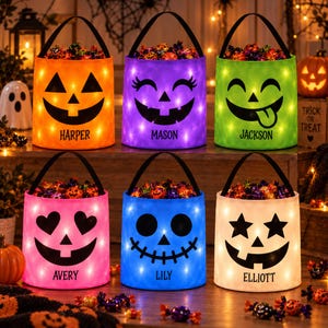 May include: Six cylindrical Halloween candy buckets with black handles and LED lights. Each bucket features a unique black face design and a personalized name: Harper, Mason, Jackson, Avery, Lily, or Elliott. The buckets are filled with candy.