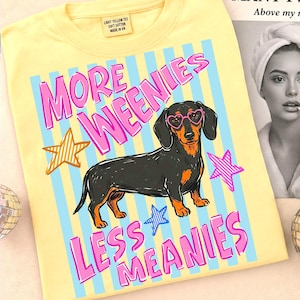 May include: Light yellow t-shirt with a graphic of a dachshund wearing heart-shaped glasses. The shirt has the text "MORE WEENIES LESS MEANIES" in pink, with stars and blue stripes. The tag reads "LIGHT YELLOW TEE SOFT COTTON MADE IN VN."