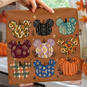 May include: A brown fabric square featuring a graphic print of nine pumpkins, each shaped with mouse-like ears. The pumpkins display various patterns including leopard print, geometric shapes, floral motifs, plaid, and celestial stars on a brown background.