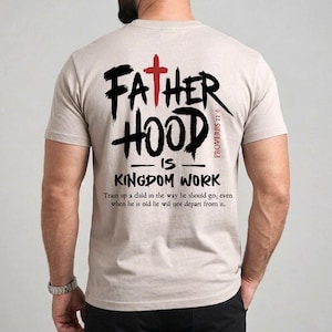 May include: Beige t-shirt with the text "FATHER HOOD IS KINGDOM WORK" in black brush lettering and a red cross. The shirt also features a Bible verse. A man is wearing the shirt.