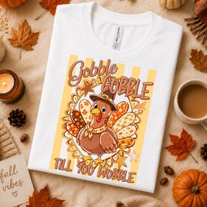 May include: A white t-shirt featuring a graphic of a brown turkey wearing a pilgrim hat and a neckerchief. The turkey is surrounded by a decorative oval frame with stars and patterned feathers. The text reads GOBBLE GOBBLE TILL YOU WOBBLE.