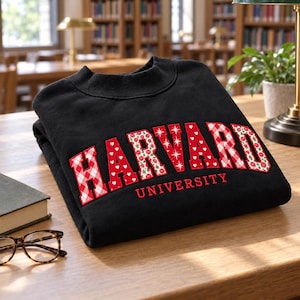 以下が含まれることがあります： 黒いクルーネックのスウェットシャツ。HARVARDの文字が、白のハート、星、花柄があしらわれた大きな赤のブロック体でデザインされており、その下にUNIVERSITYという文字が小さめの赤色で記されています。