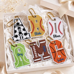 May include: Six embroidered letter keychains with gold-tone carabiners. The letters A, B, I, R, M, and G feature sports-themed patterns including soccer, baseball, football, and basketball designs, each personalized with a name in script embroidery.