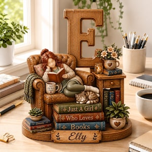 Könnte beinhalten: Eine dekorative Figur, die eine lesende Person in einem braunen Sessel vor einem großen hölzernen Buchstaben E zeigt. Der Sockel enthält gestapelte Bücher mit der Aufschrift Just A Girl Who Loves Books und Elly, eine Katze, eine Tasse und Pflanzen.