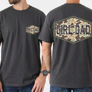 May include: Charcoal gray t-shirts with a camouflage design and the text "GIRL DAD". The front of the shirt has a small pocket with the same design. The back of the shirt has a large, hexagonal design with the text "GIRL DAD".