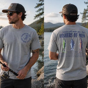 Op de afbeelding: Een lichtgrijs t-shirt met de tekst "FISHERS OF MEN" en een vishaakontwerp. De achterkant van de shirt heeft de tekst "Jesus will make you FISHERS OF MEN" met drie visillustraties. Het shirt wordt gedragen door een persoon die een hoed en zonnebril draagt.