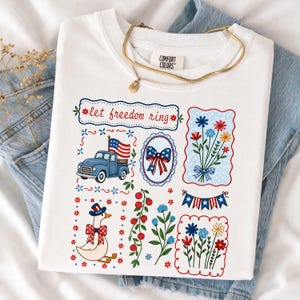 May include: White t-shirt featuring patriotic designs. The graphics include a blue truck with an American flag, floral bouquets, a duck in a patriotic hat, and the text "let freedom ring". A gold necklace is draped on the shirt.