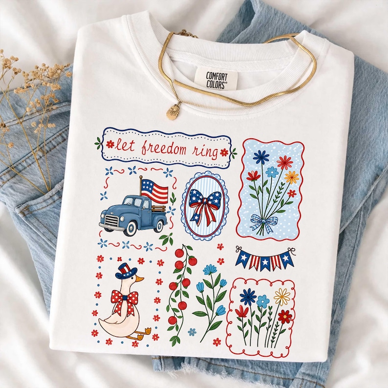 May include: White t-shirt featuring patriotic designs. The graphics include a blue truck with an American flag, floral bouquets, a duck in a patriotic hat, and the text "let freedom ring". A gold necklace is draped on the shirt.
