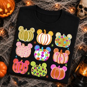 Peut inclure: Un t-shirt noir avec une impression graphique de neuf citrouilles, chacune conçue avec des formes d'oreilles de souris. Les citrouilles présentent divers motifs dont des rayures, des pois, des cœurs, des taches de léopard et des motifs floraux.