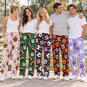 May include: Five pairs of custom pajama pants featuring sports themes. The first pair is white with baseballs, gloves, and the name Sophia 35. The second is green with footballs and the name Emma 99. The third is black with soccer balls and the name Olivia 28. The fourth is black with basketballs and the name Mason 12. The fifth is purple with volleyballs and the name Logan 23.