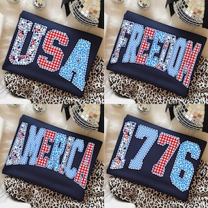 May include: Four navy blue sweatshirts with patriotic text: "USA", "FREEDOM", "AMERICA", and "1776". The letters feature red gingham, blue leopard print, and floral patterns. The sweatshirts are displayed on a leopard print fabric.