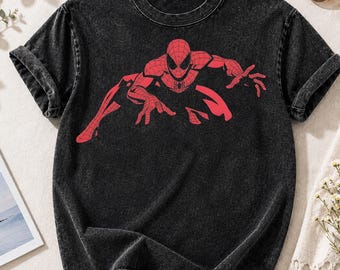 May include: A black acid-wash t-shirt featuring a red graphic print of a superhero in a crouching position. The shirt has a crew neckline and short sleeves, with the character design centered on the chest area, showcasing a web-like pattern.