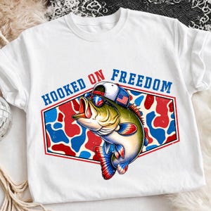 May include: White t-shirt featuring a cartoon bass wearing sunglasses and a hat with an American flag design. The shirt has the text "HOOKED ON FREEDOM" in blue. The fish is set against a red, white, and blue camouflage pattern.