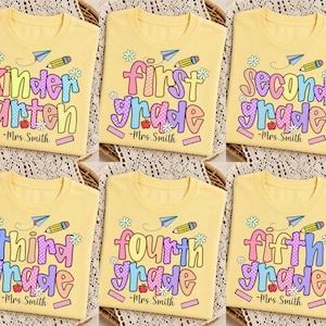 Pode incluir: Uma t-shirt amarelo-claro com um gráfico colorido de temática escolar, incluindo um avião de papel, um lápis, margaridas e uma maçã. O texto apresenta vários níveis escolares e o nome Mrs. Smith numa fonte lúdica e multicolorida.