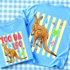 Op de afbeelding: Twee lichtblauwe t-shirts met kangoeroe-afbeeldingen. Eén shirt heeft de tekst "TOO DA LOO" in rood en roze. Beide shirts tonen een kangoeroe, gele strepen en kleurrijke sterren.