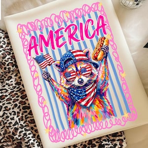 May include: A cream-colored tote bag featuring a colorful raccoon wearing patriotic sunglasses, a hat, and a bandana. The raccoon is holding a hot dog and an American flag. The word "AMERICA" is in pink, with a pink border and blue stripes.