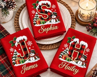May include: Three red folded kitchen towels featuring a large letter S, J, or H in a red and black buffalo plaid pattern, topped with snow, holly, and a snowman. Each towel is personalized with the names Sophia, James, or Hailey in gold script.
