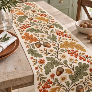 May include: A long rectangular table runner featuring a repeating botanical pattern of green and orange oak leaves, brown acorns, small white flowers, and clusters of red berries printed on a cream-coloured fabric background.