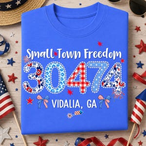 May include: A blue t-shirt featuring the text "Small Town Freedom" and the numbers "30474" in a patriotic style. The text "VIDALIA, GA" is also present. The design includes stars, bows, and firework illustrations.