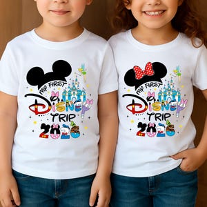 May include: Two white t-shirts with a colourful "My First Disney Trip 2020" graphic. One shirt features Mickey Mouse ears, the other Minnie Mouse ears with a red polka dot bow. Both shirts have a castle and fairy design.