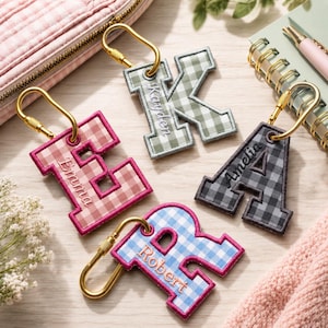 May include: Four embroidered letter keychains with gold-tone carabiners. The letters E, K, A, and R feature gingham patterns in pink, green, black, and blue. Each letter is personalized with stitched names: Emma, Kayden, Amelia, and Robert.