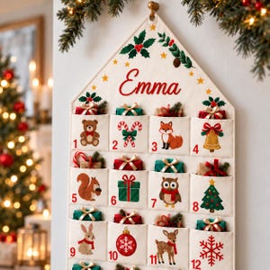 Könnte beinhalten: Ein cremefarbener Stoff-Adventskalender in Hausform mit dem in Rot gedruckten Namen Emma. Er verfügt über zwölf rot nummerierte Taschen, die mit Illustrationen von Tieren, Geschenken, Zuckerstangen und weihnachtlichen Motiven verziert sind.