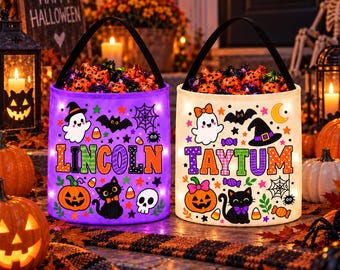 May include: Two cylindrical Halloween candy buckets with black handles, one purple and one white. Both feature colorful prints of ghosts, pumpkins, cats, bats, and spiderwebs. The purple bucket displays the name LINCOLN, and the white bucket displays TAYTUM.