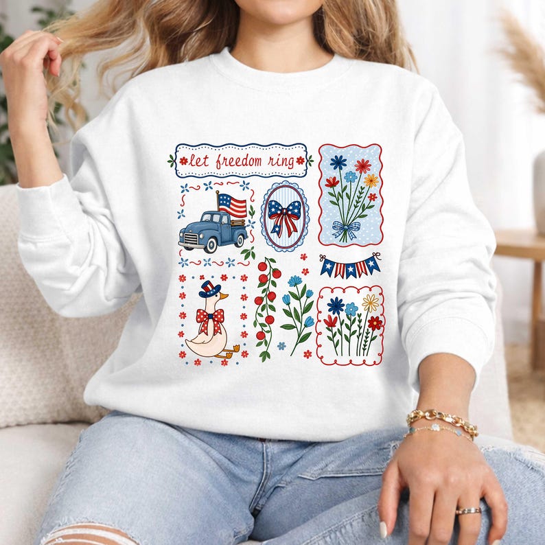 May include: White sweatshirt with patriotic designs. It features a blue truck with a flag, floral arrangements, a goose in a patriotic hat, and the text "let freedom ring".