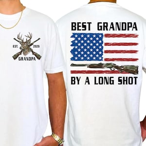 Puede incluir: Camiseta blanca con un ciervo y rifles cruzados, el texto "EST. 2026 GRANDPA" en la parte delantera. La parte trasera presenta un diseño de bandera americana con un rifle y el texto "BEST GRANDPA BY A LONG SHOT."