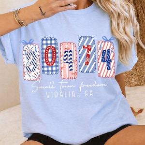 May include: Light blue t-shirt with a patriotic design. The shirt features the numbers "30474" in red, white, and blue patterns, with the words "Small Town freedom VIDALIA, GA" in white script.